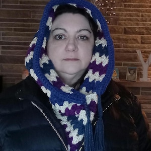 May include: A handmade crochet hood and scarf set in a geometric pattern. The hood and scarf feature a combination of blue, purple, and cream colors. The scarf has tassels at the ends. The person in the image is wearing a black jacket.