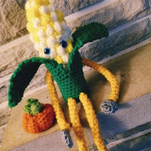 May include: A crocheted corn on the cob with a green husk and yellow kernels. The corn has a pair of blue eyes and is sitting on a stone surface with a crocheted orange pumpkin.