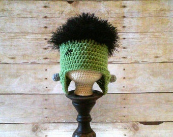 May include: Green and black crocheted hat with a black fuzzy top and gray ear flaps. The hat is on a wooden stand.