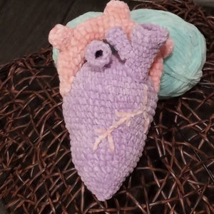 May include: A crocheted heart shaped plush toy in shades of purple, pink, and light blue. The heart is sitting on a woven brown basket.