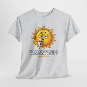 May include: Light gray t-shirt featuring a tired sun cartoon character holding a coffee cup. The sun is yellow with orange rays and a sleepy expression. Text on the shirt reads "CURRENTLY OPERATING AT 12% LUMINOSITY."