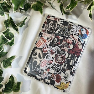 Puede incluir: Una funda transparente para teléfono con un patrón de cuadros blanco y negro, cubierta de pegatinas con temática de películas de terror. Las pegatinas presentan imágenes de personajes de terror icónicos y frases como "HORROR MOVIE ADDICT" y "FRIDAY THE 13TH".