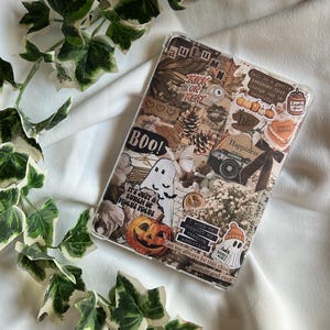 Puede incluir: Funda transparente para teléfono decorada con pegatinas de temática otoñal, incluyendo calabazas, fantasmas y el texto "BOO!". El diseño incorpora hojas de otoño, una cámara y las palabras "Trick or Treat".