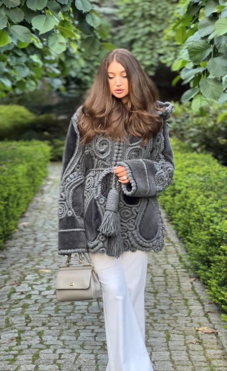 May include: A gray, intricately designed coat with a hood and decorative tassels. The coat features detailed patterns and is paired with white pants and a gray handbag. The image is set outdoors with a stone path and greenery.