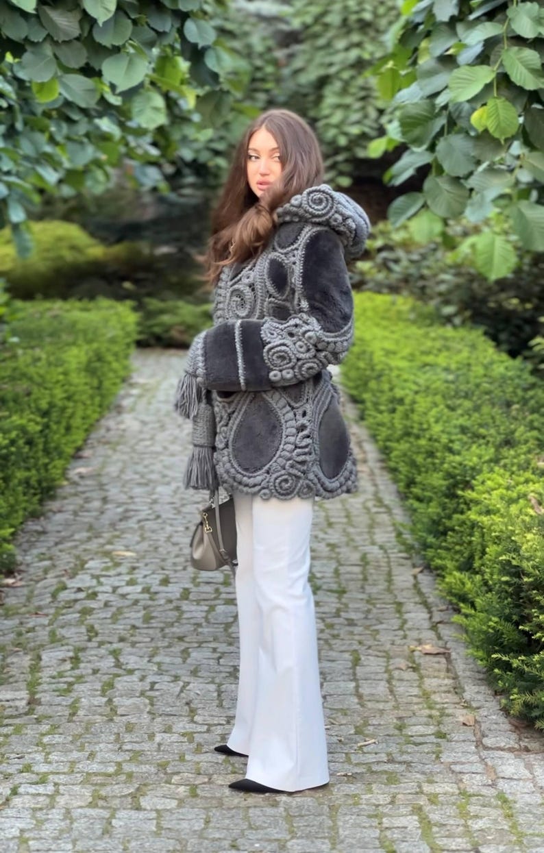 May include: A gray hooded coat with intricate, decorative stitching and a matching belt. The coat features a unique design with a combination of textures and patterns. The person is wearing white pants and carrying a gray purse.