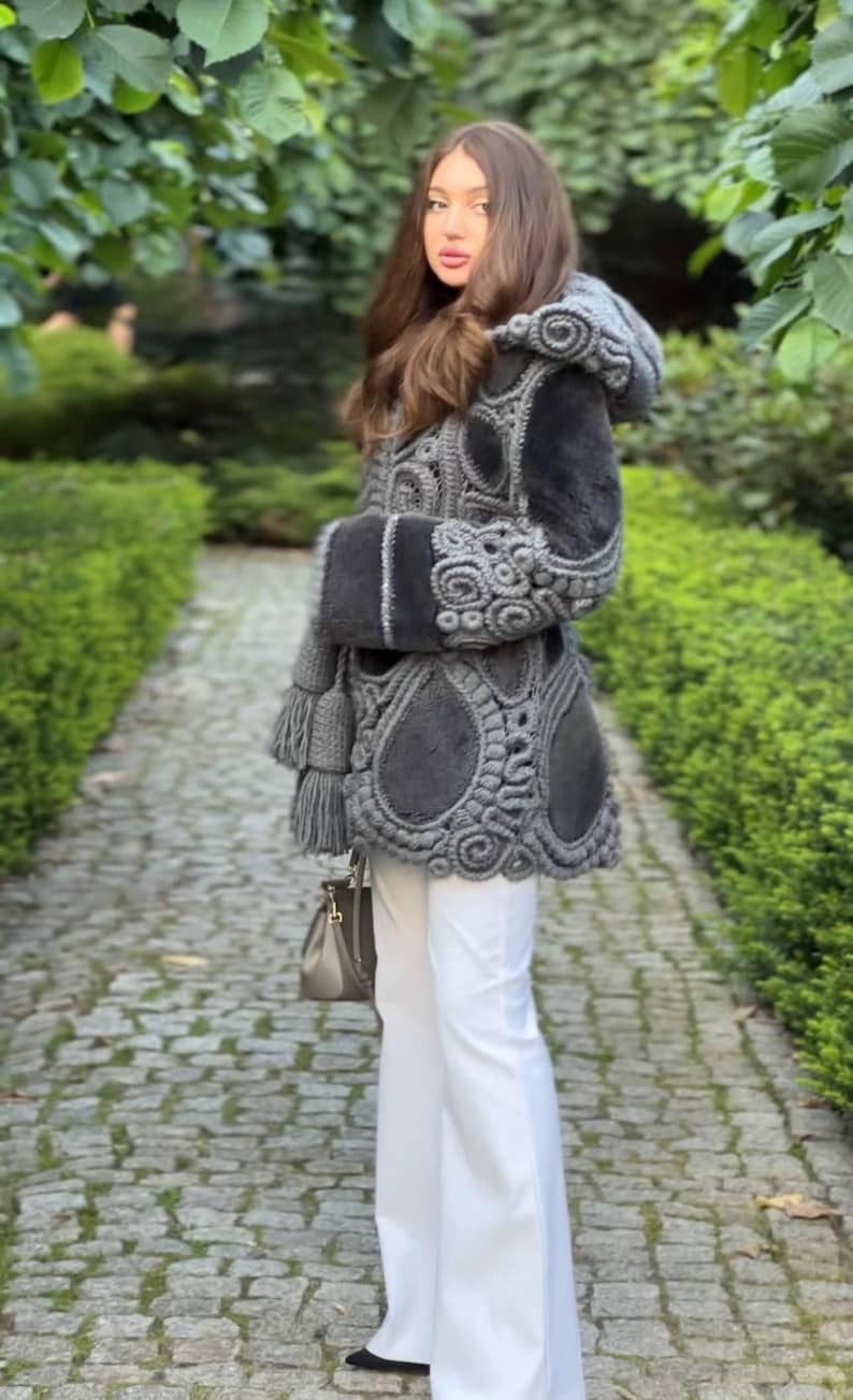 May include: A gray hooded coat with intricate crochet detailing and a fur-lined interior. The coat features a belt, tassels, and is paired with white pants and a gray handbag. The coat is a unique fashion statement.