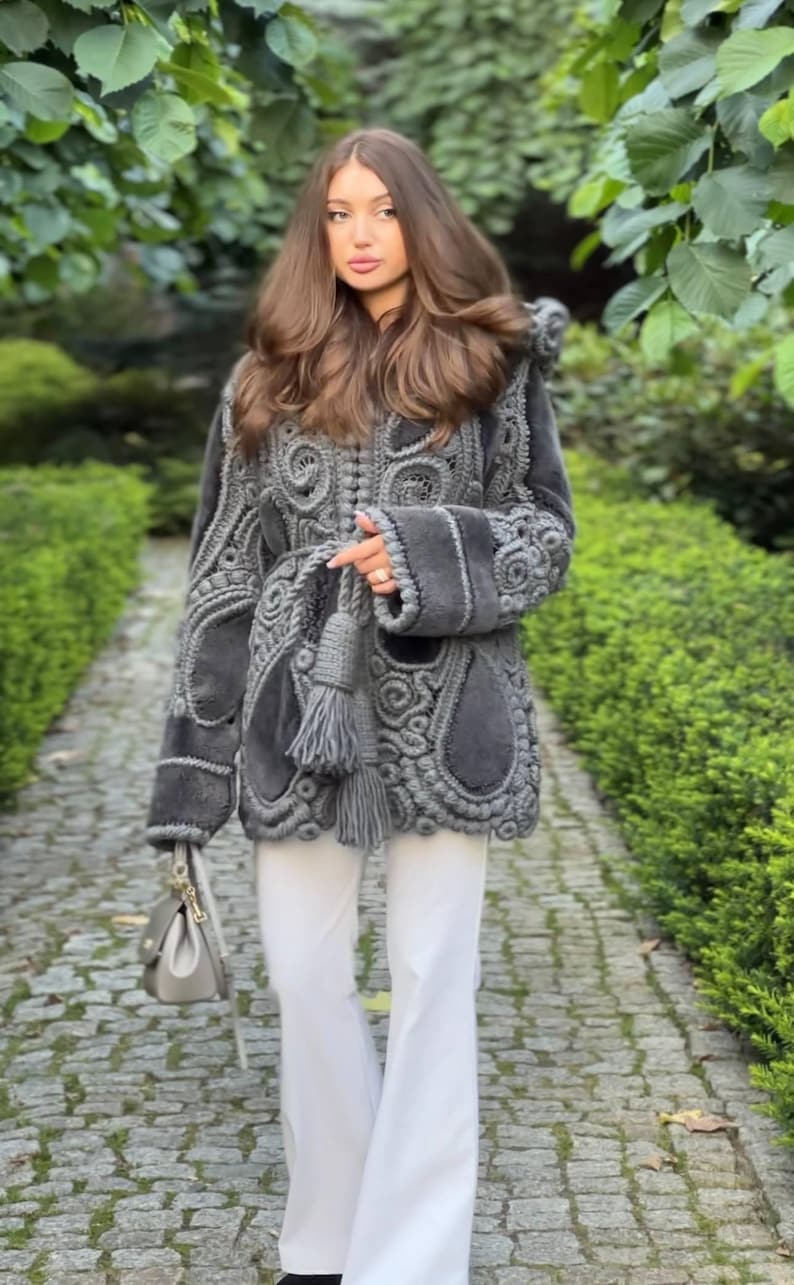 May include: A gray, intricately designed coat with a unique, textured pattern. The coat features a detailed, openwork design and is paired with white pants. A small gray handbag and a tassel are also visible.