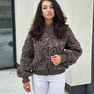 May include: A brown, zip-up knitted jacket with a distinctive textured pattern. The jacket has a high collar and elaborate, swirling designs across the front and sleeves. The sleeves are balloon-shaped. The jacket is made of a soft, knitted material.