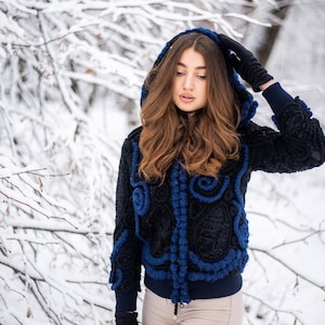 May include: A black hooded jacket with blue accents and intricate detailing. The jacket features a textured design and is worn with black gloves and tan pants. The background shows snow-covered trees, suggesting a winter setting.
