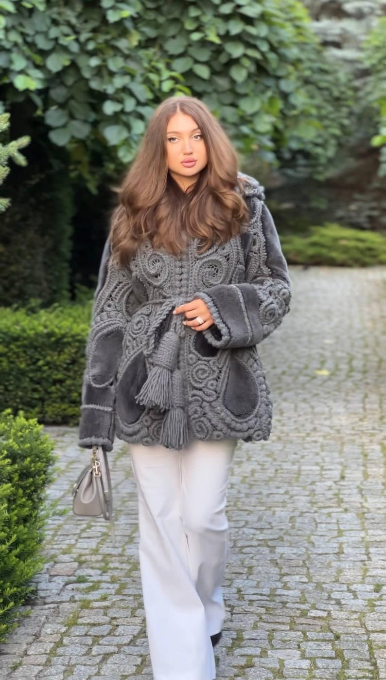 May include: A gray coat with intricate crochet detailing and a hood. The coat features a unique design with textured patterns and a matching tassel belt. The person is wearing white pants and carrying a gray handbag.