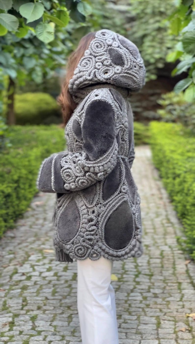 May include: A gray coat with a hood, featuring intricate crochet detailing. The coat has a plush, textured appearance with a tailored fit. The design includes floral and teardrop-shaped patterns, creating a unique and elegant look.