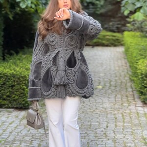 May include: A gray, intricately designed coat with a unique pattern, paired with white pants and a gray handbag. The coat features a textured, openwork design with a belted waist and fringe detailing. The outfit is worn outdoors.
