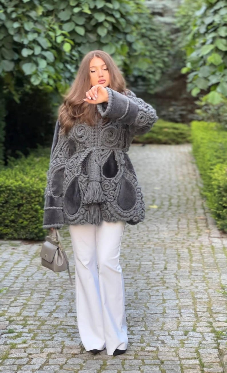 May include: A gray, intricately designed coat with a unique pattern, paired with white pants and a gray handbag. The coat features a textured, openwork design with a belted waist and fringe detailing. The outfit is worn outdoors.