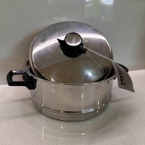 May include: A stainless steel cooking pot with a lid, black handles and a black knob. A tag is attached to the pot. The pot is on a white countertop.