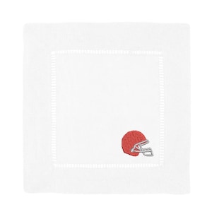 May include: A square white linen napkin featuring a decorative hemstitched border and a small, detailed embroidery of a solid orange football helmet with a silver face mask positioned in the lower right corner of the fabric square.