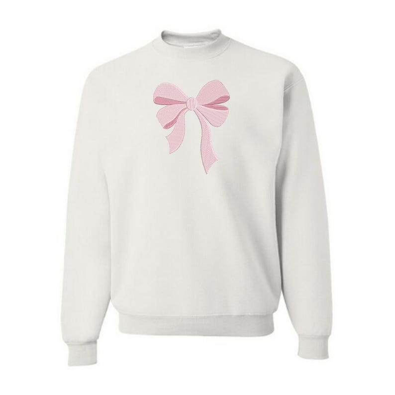 Peut inclure: Un sweat-shirt blanc &agrave; manches longues et col rond, orn&eacute; d'un n&oelig;ud rose clair brod&eacute; au centre. Le v&ecirc;tement pr&eacute;sente un col, des poignets et un ourlet c&ocirc;tel&eacute;s, offrant une apparence nette et structur&eacute;e avec un &eacute;l&eacute;ment d&eacute;coratif central.