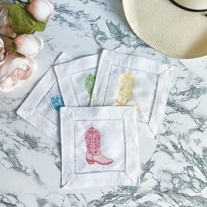 May include: Four white linen napkins with embroidered cowboy boot designs. The boots are in different colors: red, yellow, blue, and green.