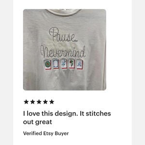 May include: A light gray long-sleeve shirt with the embroidered text "Pause... Nevermind". Below the text are embroidered images of Mahjong tiles. The shirt has a crew neck.