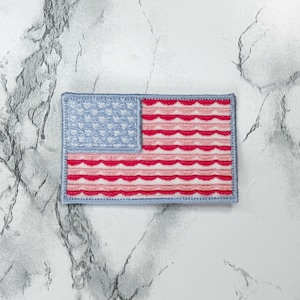 May include: A rectangular embroidered patch featuring a stylized American flag design. The canton is light blue with white heart-shaped patterns, while the stripes consist of alternating wavy lines in shades of red and white, all bordered by light blue.