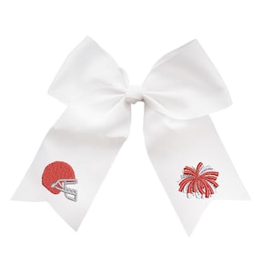 May include: A large white hair bow made of stiff ribbon with two long tails. The left tail features an embroidered red football helmet with a silver facemask, and the right tail features an embroidered red and silver cheerleader pom-pom.