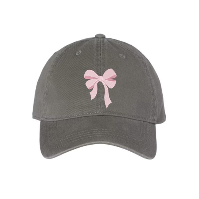 Peut inclure: Une casquette de baseball gris anthracite orn&eacute;e d'un n&oelig;ud rose clair brod&eacute; au centre du panneau avant. La casquette pr&eacute;sente une calotte structur&eacute;e, une visi&egrave;re incurv&eacute;e et des &oelig;illets cousus pour la ventilation, offrant un design &eacute;pur&eacute;.