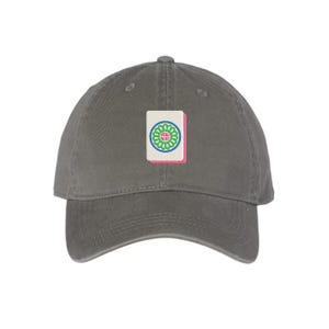 May include: A gray baseball cap with an embroidered patch featuring a Mahjong tile. The tile has a white background with a green and blue floral design and a pink border. The cap is made of a soft fabric.