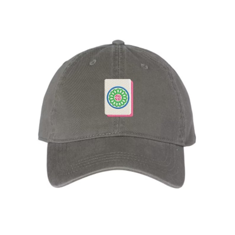May include: A gray baseball cap with an embroidered patch featuring a Mahjong tile. The tile has a white background with a green and blue floral design and a pink border. The cap is made of a soft fabric.