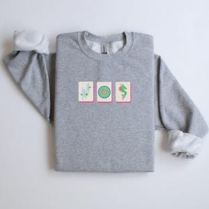 May include: A heather gray sweatshirt featuring three embroidered Mahjong tile designs. The tiles are rectangular with pink borders and depict bamboo, a green circle, and a green dragon. The sleeves have white cuffs.