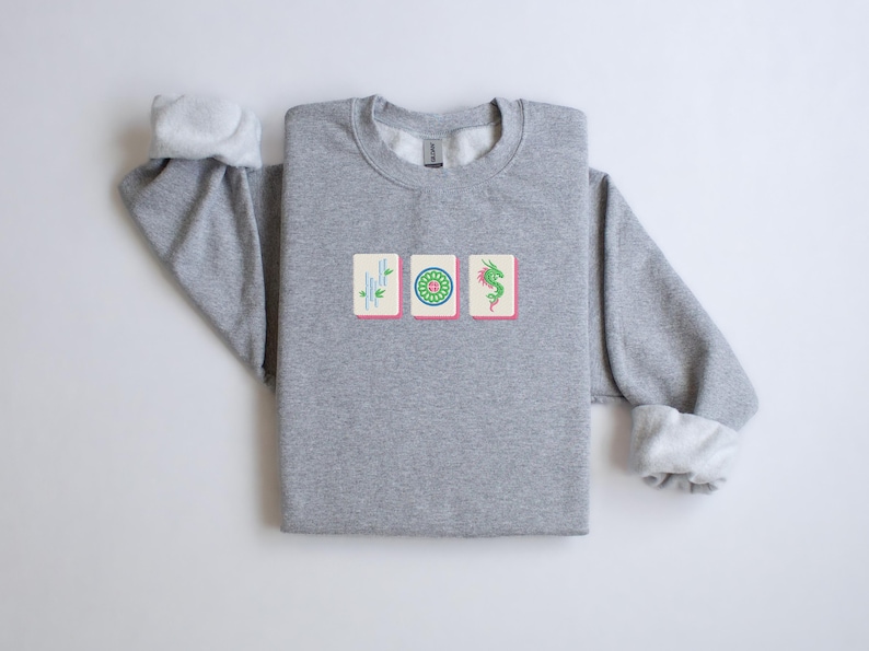 May include: A heather gray sweatshirt featuring three embroidered Mahjong tile designs. The tiles are rectangular with pink borders and depict bamboo, a green circle, and a green dragon. The sleeves have white cuffs.