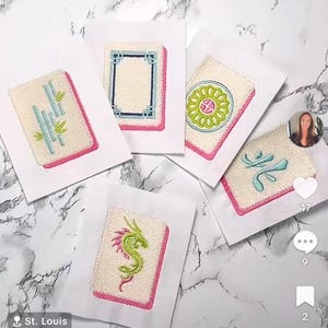 May include: Five embroidered Mahjong tile designs on white fabric, each with a different symbol and a pink border. The tiles are arranged on a marble surface. The text "CreativelyEmb" and "St. Louis" are visible.