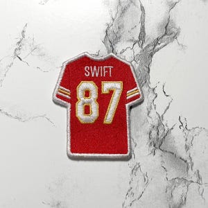 May include: Red and gold embroidered jersey patch with the name "SWIFT" above the number "87". The patch has a white border and is designed to resemble a football jersey.