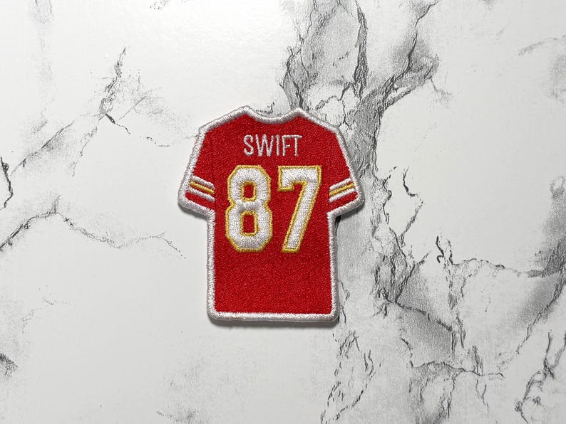 May include: Red and gold embroidered jersey patch with the name "SWIFT" above the number "87". The patch has a white border and is designed to resemble a football jersey.