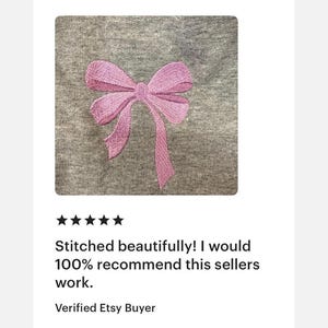 Peut inclure: Un v&ecirc;tement en tissu gris chin&eacute; arborant un motif de n&oelig;ud rose clair brod&eacute; au centre. Le n&oelig;ud pr&eacute;sente des motifs de points visibles qui cr&eacute;ent de la texture et de la profondeur sur le mat&eacute;riau de fond gris aux tons neutres et doux.
