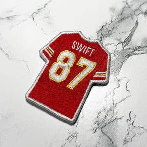 May include: Red and gold embroidered patch in the shape of a football jersey. The jersey patch features the name "SWIFT" above the number "87" in white and gold. The patch has a white border.