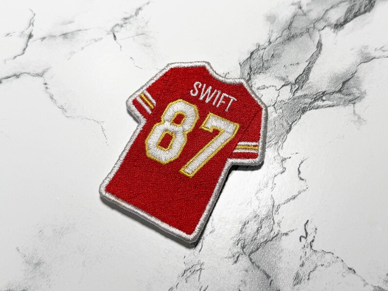 May include: Red and gold embroidered patch in the shape of a football jersey. The jersey patch features the name "SWIFT" above the number "87" in white and gold. The patch has a white border.