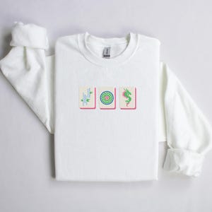 May include: White crew neck sweatshirt featuring three embroidered Mahjong tiles. The tiles are in a row, with a bamboo, a flower, and a green dragon design. The sweatshirt has long sleeves with rolled cuffs.