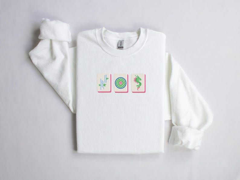 May include: White crew neck sweatshirt featuring three embroidered Mahjong tiles. The tiles are in a row, with a bamboo, a flower, and a green dragon design. The sweatshirt has long sleeves with rolled cuffs.