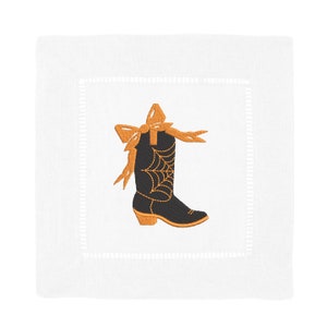Puede incluir: Una servilleta de c&oacute;ctel de lino blanco con una bota vaquera negra bordada con un lazo naranja y un dise&ntilde;o de telara&ntilde;a.