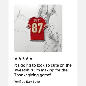 May include: Red and gold jersey patch with the name "SWIFT" and the number "87". The patch is designed to resemble a football jersey. The background is a white and gray marble pattern.