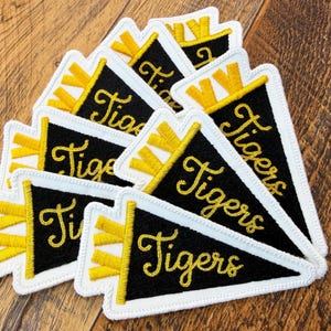 May include: A collection of black pennant patches with the word "Tigers" in gold script. Each patch has a white border and yellow accents. The patches are arranged on a wooden surface.