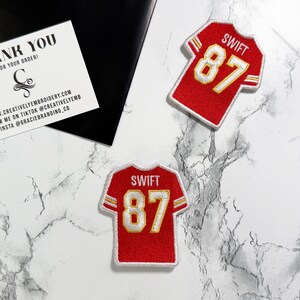 May include: Two red jersey patches with the name "SWIFT" and the number "87" in gold. A white card with black text that reads "THANK YOU" and the website address. The patches are on a marble surface.
