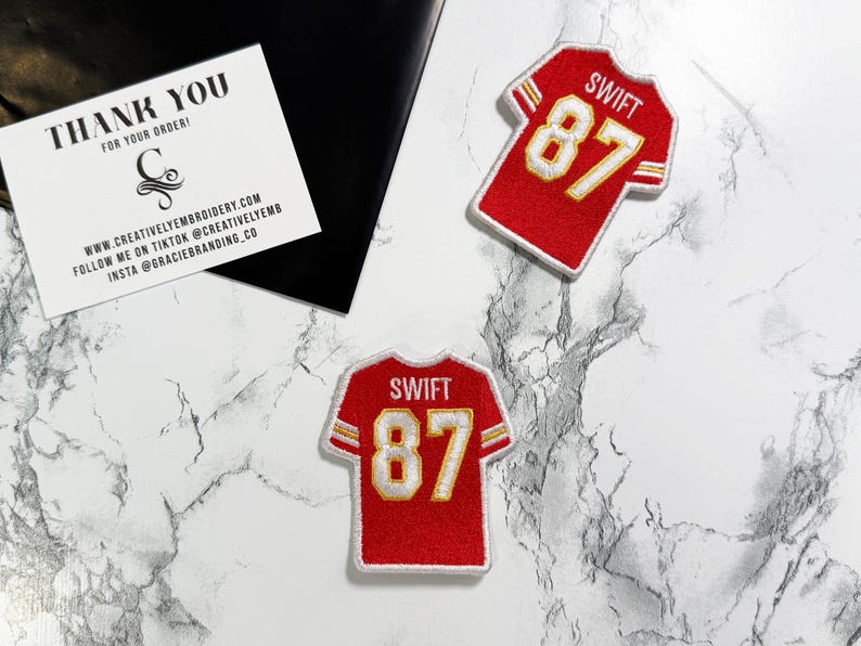 May include: Two red jersey patches with the name "SWIFT" and the number "87" in gold. A white card with black text that reads "THANK YOU" and the website address. The patches are on a marble surface.