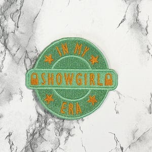 May include: A green embroidered patch with the words "IN MY SHOWGIRL ERA" in orange thread. The patch is circular with a banner across the middle. Four orange stars are embroidered around the text. The patch is on a marble background.