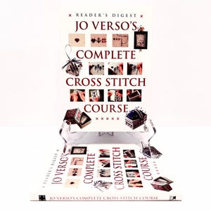 May include: A Reader's Digest book titled "Jo Verso's Complete Cross Stitch Course" is displayed on a clear stand. The cover features cross-stitch examples and instructional images. The book is white with red and black text.