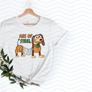 May include: White t-shirt featuring a cartoon illustration of Slinky Dog with the text "ABS OF STEEL" in orange and green. The design showcases the character's spring body and friendly expression, perfect for fans of the animated film.