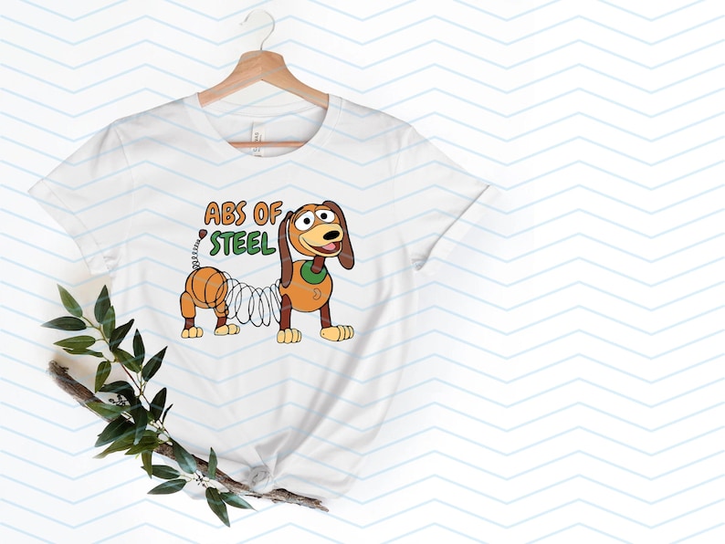 May include: White t-shirt featuring a cartoon illustration of Slinky Dog with the text "ABS OF STEEL" in orange and green. The design showcases the character's spring body and friendly expression, perfect for fans of the animated film.