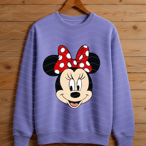 May include: A lavender sweatshirt featuring a Minnie Mouse graphic. The graphic shows Minnie's face with black ears, a red bow with white polka dots, and a smiling expression. The sweatshirt is hanging on a wooden hanger.