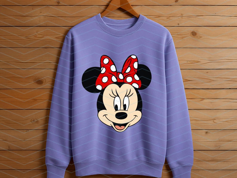 May include: A lavender sweatshirt featuring a Minnie Mouse graphic. The graphic shows Minnie's face with black ears, a red bow with white polka dots, and a smiling expression. The sweatshirt is hanging on a wooden hanger.