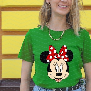 May include: Green t-shirt featuring a Minnie Mouse graphic. The graphic shows Minnie's face with black ears, a red bow with white polka dots, and a smiling expression. The shirt is made of a soft material.