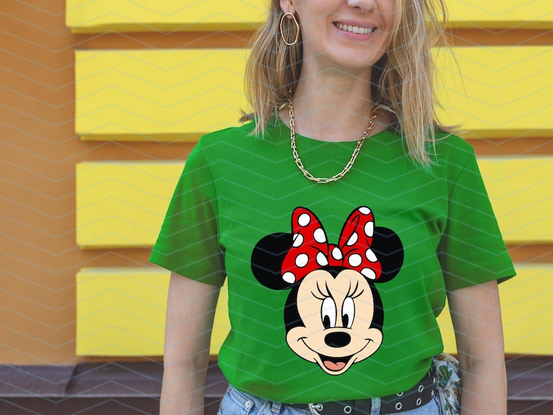 May include: Green t-shirt featuring a Minnie Mouse graphic. The graphic shows Minnie's face with black ears, a red bow with white polka dots, and a smiling expression. The shirt is made of a soft material.